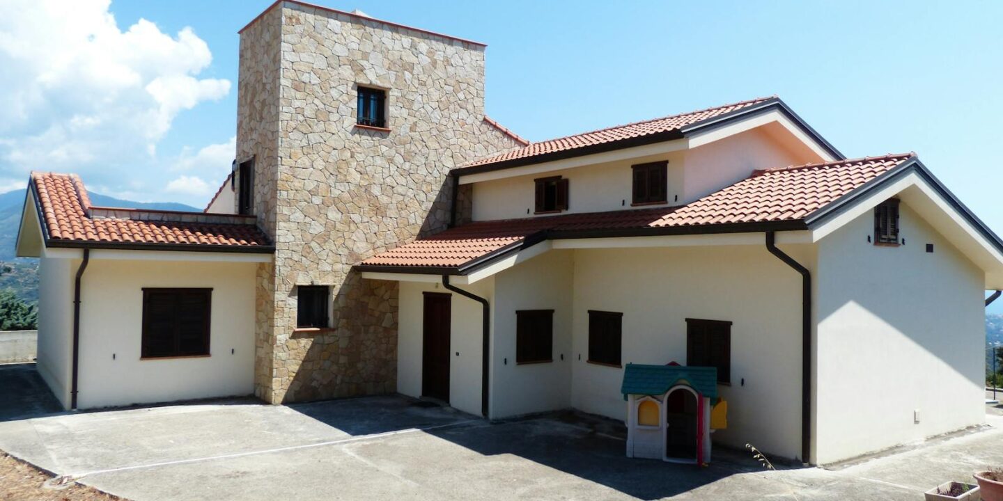 Spacious family house featuring a stone facade and red tile roof, set outdoors under a bright blue sky.
