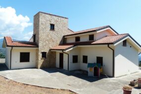 Spacious family house featuring a stone facade and red tile roof, set outdoors under a bright blue sky.