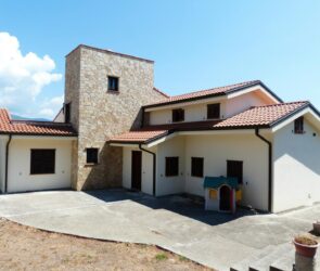 Spacious family house featuring a stone facade and red tile roof, set outdoors under a bright blue sky.
